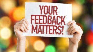 A man’s hands hold a sign that reads “ Your Feedback Matters!” against a background of multicolored lights.