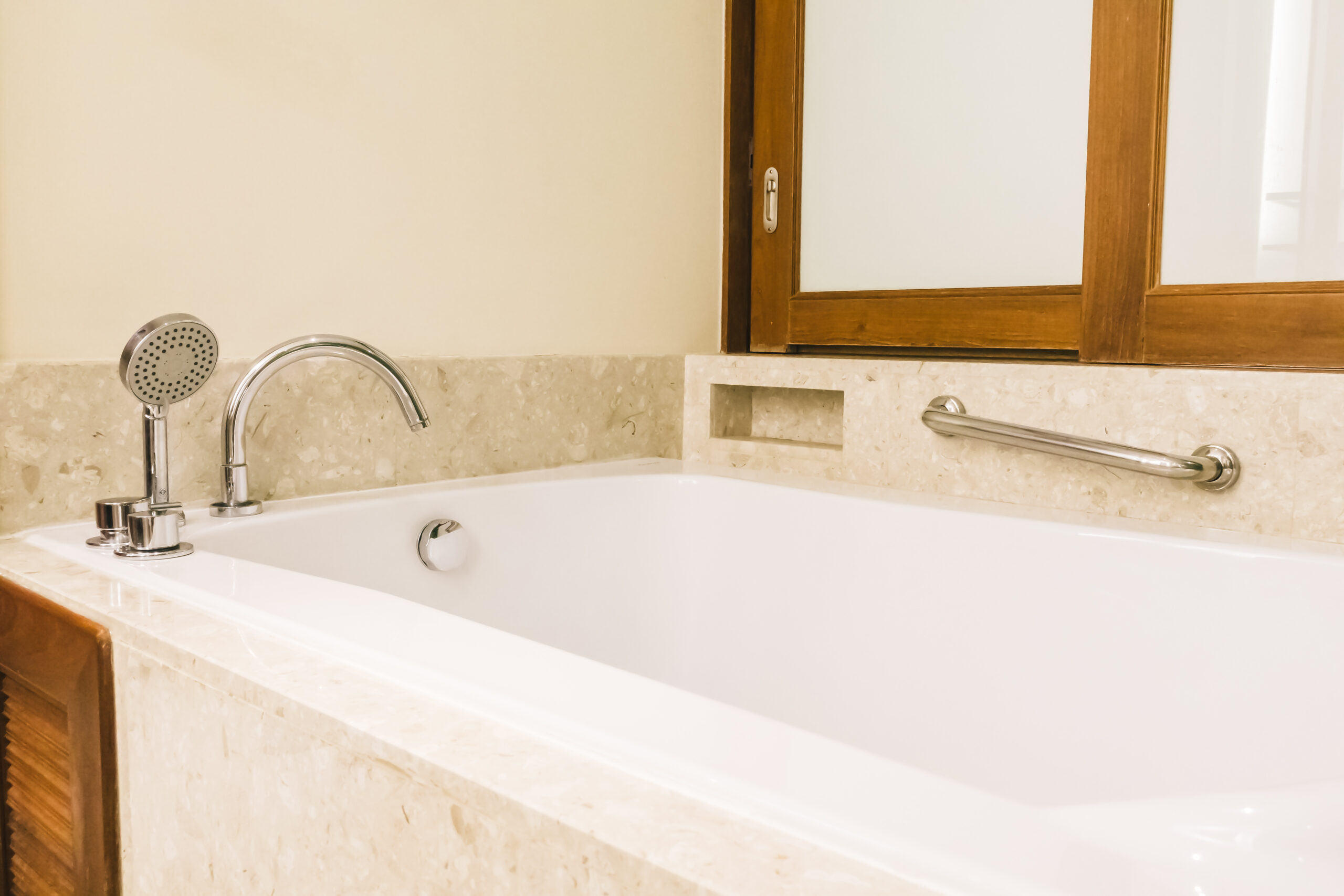This image features a beautiful rectangular bathtub with a surrounding ledge. There is a grab bar on the wall along with a mirror the homeowner put up.